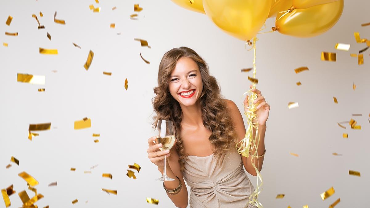 attractive young stylish woman celebrating new year, drinking champagne holding air balloons, golden confetti flying, smiling happy, white background, isolated, wearing party dress