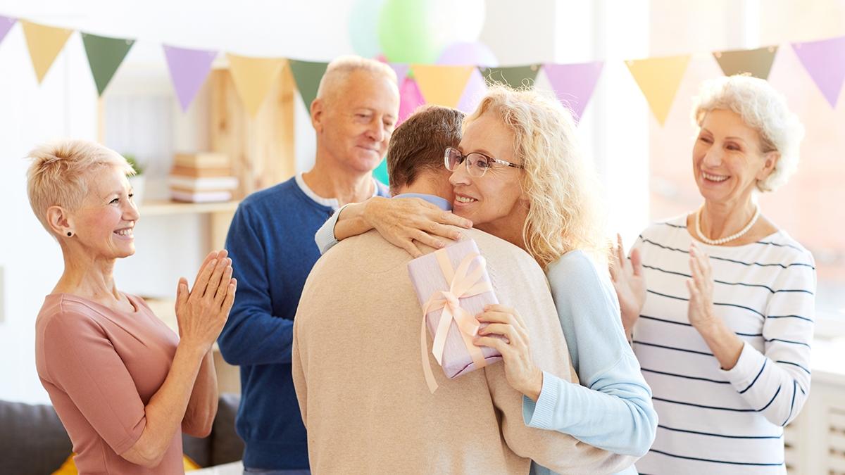 Positive attractive mature lady in glasses standing in living room decorated with garland and hugging friend at birthday party while excited women and man clapping her during congratulations.