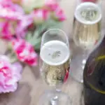 Bottle of Prosecco and two champagne glasses on a rustic garden picnic table with blossom or flowers in the background. Springtime or Summertime.