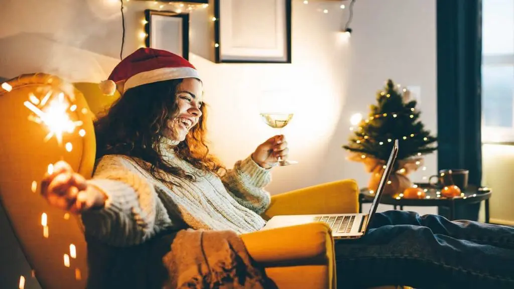 Happy young woman wearing Santa hat, drinking champagne, talking with friends on video call celebrate New Year party.