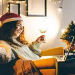 Happy young woman wearing Santa hat, drinking champagne, talking with friends on video call celebrate New Year party.