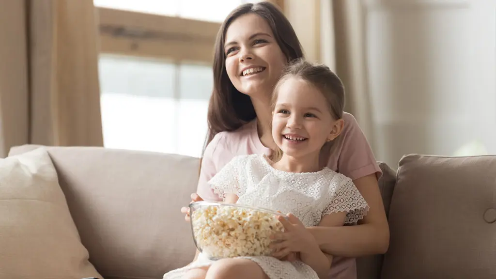 Celebrate Mom this Mother’s Day with a Movie Marathon: Here’s How Happy mom and girl laughing watching tv on sofa