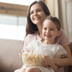 Happy mom and girl laughing watching tv on sofa