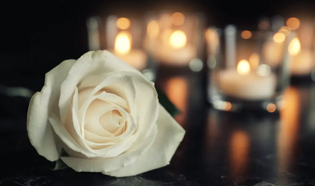 White rose and blurred burning candles on table in darkness, space for text. Funeral symbol