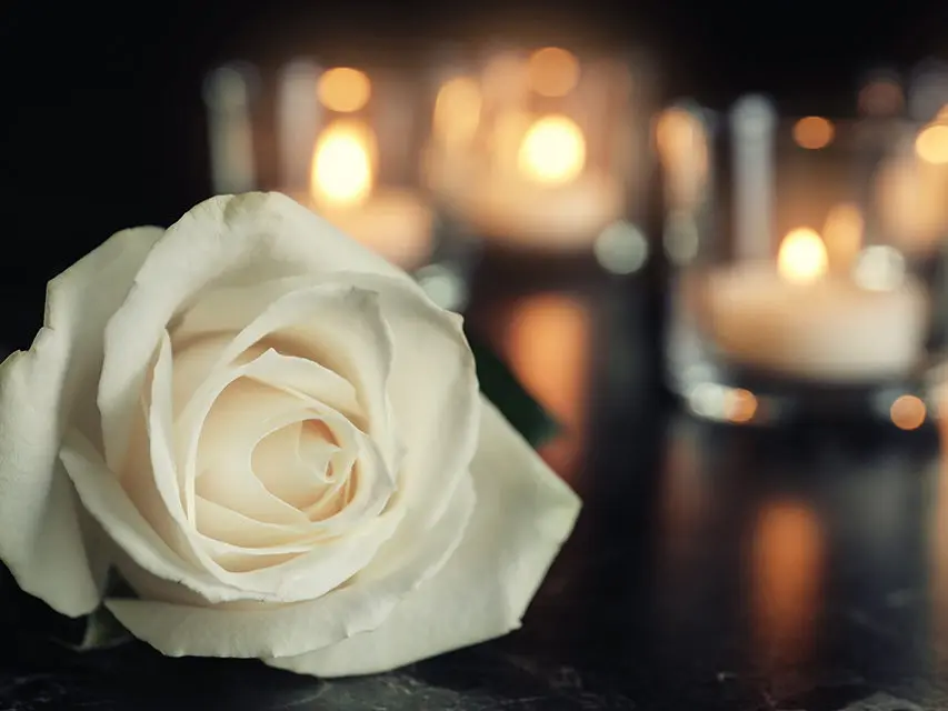White rose and blurred burning candles on table in darkness, space for text. Funeral symbol