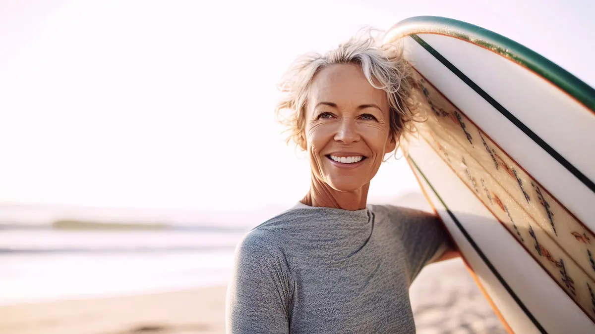 Middle aged woman holding a surfboard on a beach, radiating vita