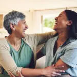 Funny, senior mother and hug daughter in home, bonding and laughing together. Happy, elderly mom and embrace woman, relax and smile with care, love and enjoying time on living room sofa with family.